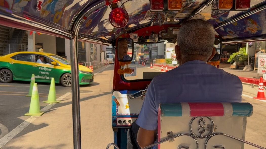 View from inside a colorful tuk-tuk in Bangkok traffic, with the driver visible from behind and a yellow-green taxi beside them on a busy city street in Thailand