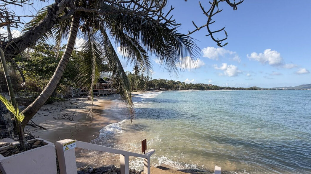 Quiet sandy beach at Chaweng Noi, Koh Samui, with leaning palm trees, gentle turquoise waves, and a traditional Thai building visible in the background