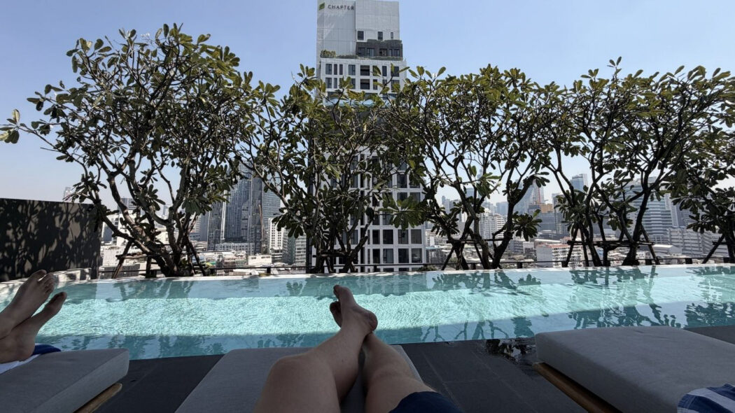 Rooftop infinity pool at Dusit D2 Samyan hotel in Bangkok, with legs resting on a sun lounger and the Bangkok city skyline visible through tropical frangipani trees