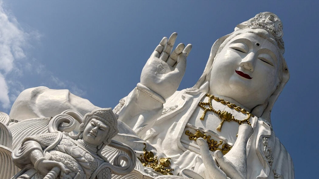 Giant white Guanyin statue in Northern Thailand with gold ornaments, hand raised in blessing gesture, photographed from below against a blue sky