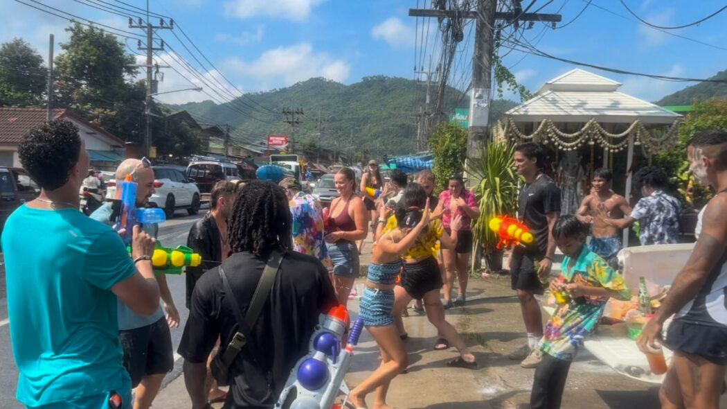 Large crowd of locals and tourists celebrating Songkran water festival on the streets of Koh Samui with water guns, hills in background, 2026