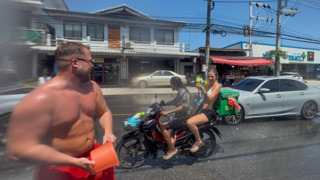 Man with orange bucket laughing during Songkran water fight on the streets of Koh Samui, Thailand 2026, with motorbike rider in background