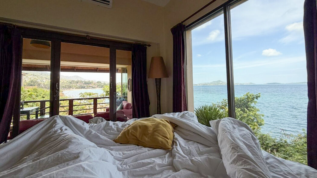 Unmade white bed in a bungalow with floor-to-ceiling windows overlooking a turquoise sea and tropical coastline in Koh Samui, Thailand