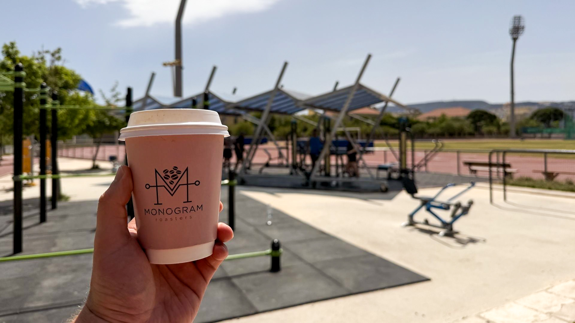 Monogram Roasters coffee cup held up in front of Chania sports stadium outdoor gym Monogram Roasters coffee cup held up in front of Chania sports stadium outdoor gym