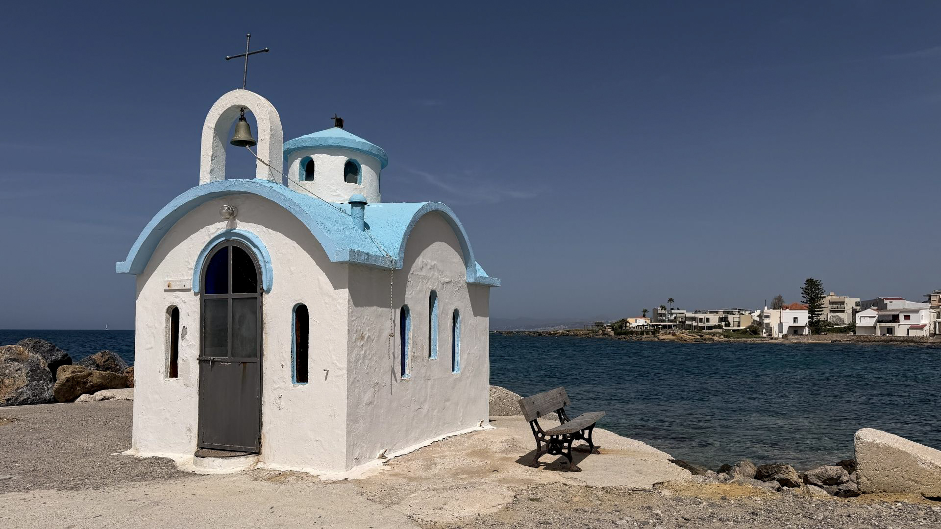 Small white and blue Greek Orthodox chapel by the sea in Crete with a wooden bench